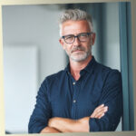 A professional portrait of Will McDonald, a personal development writer with grey hair and glasses, leaning against a wall with crossed arms in a bright office.