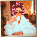 A glamorous woman in a white fluffy bathrobe and oversized jeweled sunglasses, with her hair in large pink rollers, holding a white teacup.